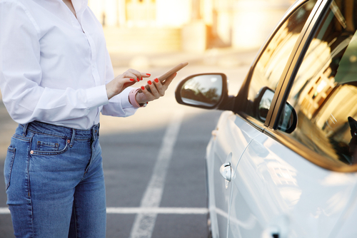 The left side of a parked white car is shown on the right, with a person on the left wearing jeans and a white, long-sleeved shirt, facing the car, smartphone in hand.