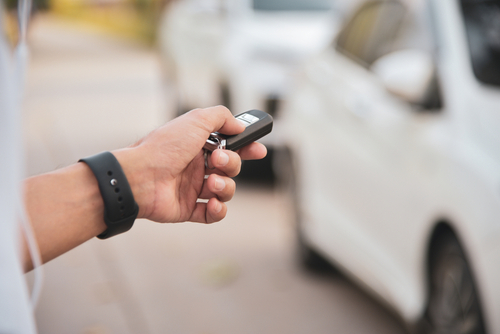 A left hand with a black wristband holds a keyless entry remote for a car. Two white cars are blurry in the background.