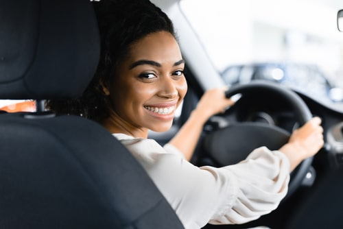 An over-the-shoulder photo of a driver, and she is turned around looking at the camera with a smiling. Her hands on the steering wheel.