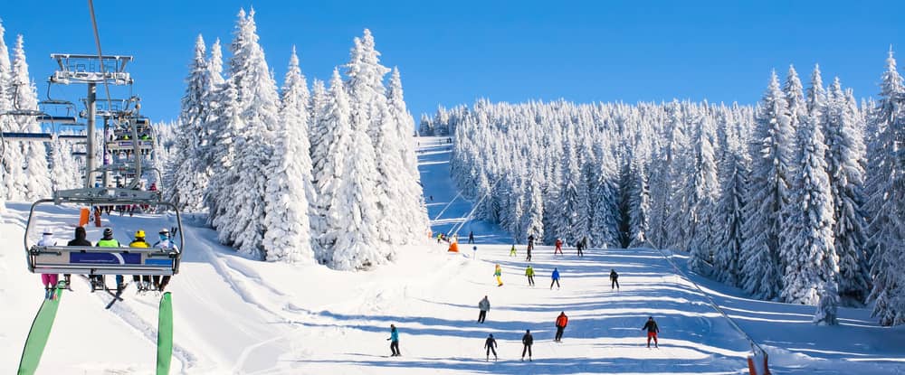 People ski on a snow-covered slope with pine trees, while a ski lift carries skiers to the left.