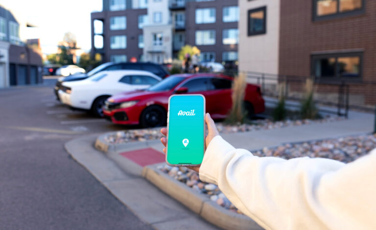 A hand holds a cell phone showing the Avail logo in a parking lot full of parked cars.