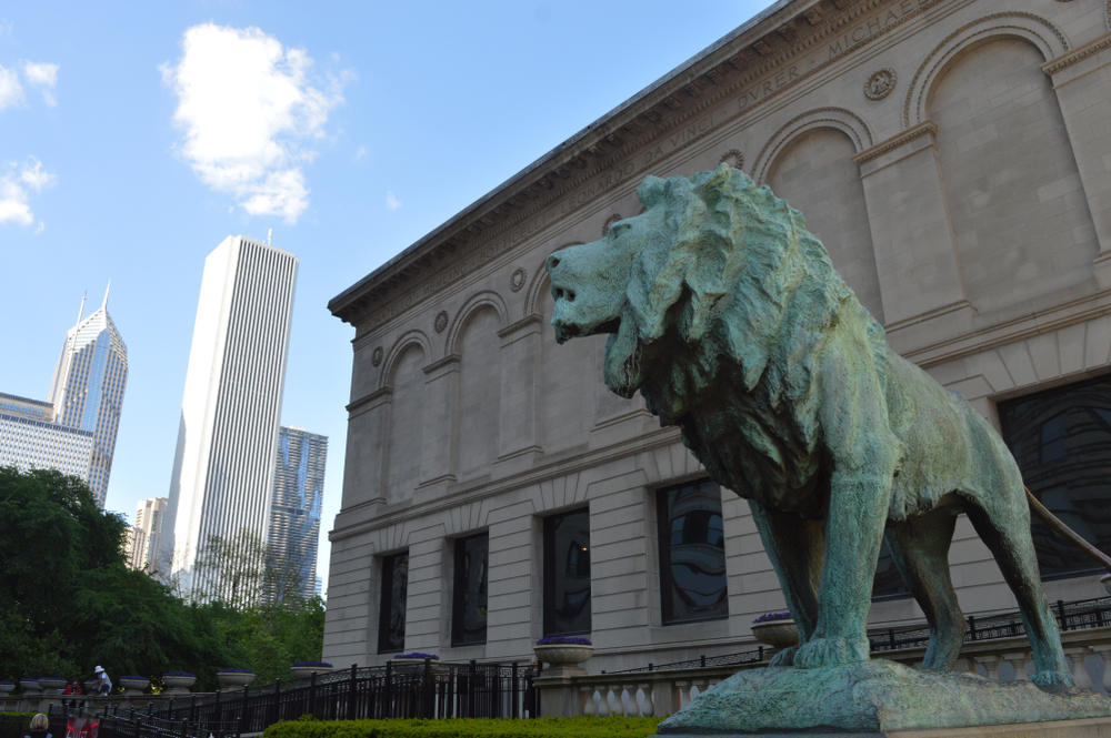 A bronze lion sculpture in front of the Art Institute of Chicago building, with skyscrapers in the background.