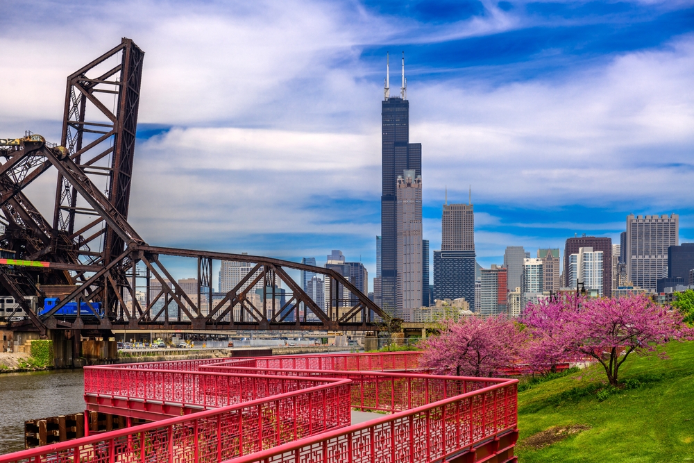 A red-railed walkway runs alongside a green park with pink blossoming trees. A bridge and skyscrapers are in the background.