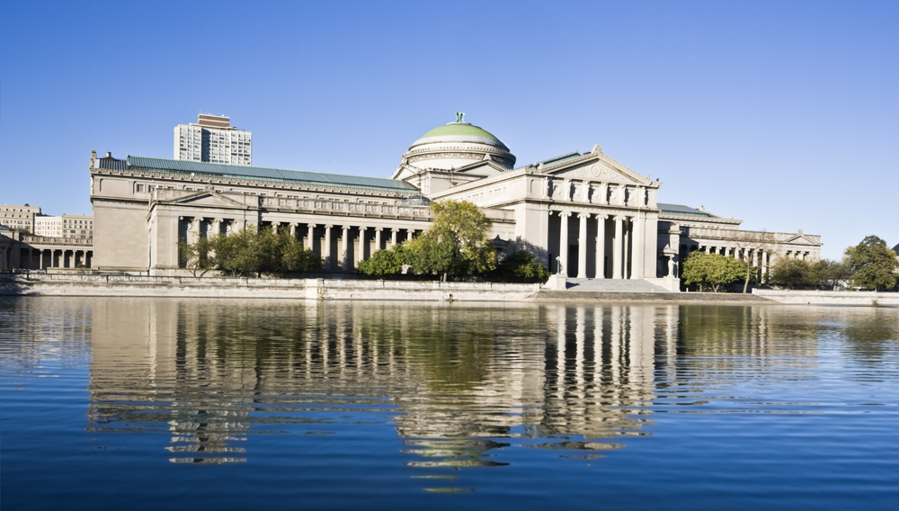 A view from across the water of the Museum of Science and Industry, with a colonnade facade and domed roof in the middle.