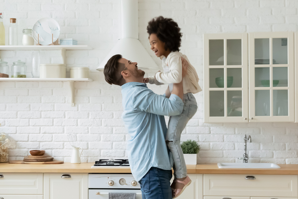 Smiling man lifts laughing young girl into the air in kitchen