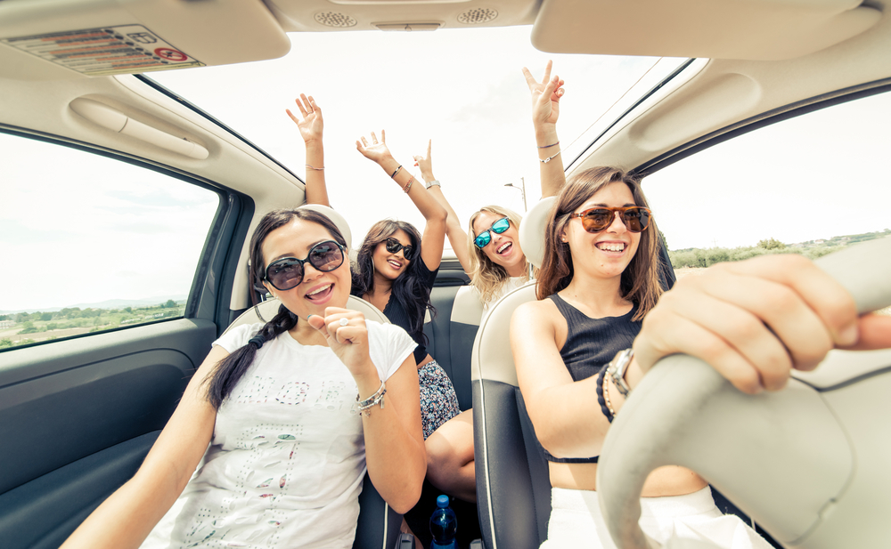 Four girls sitting in a moving vehicle. The passenger is mimicking singing into a microphone while the two in the back seat have their hands up through the sunroof.