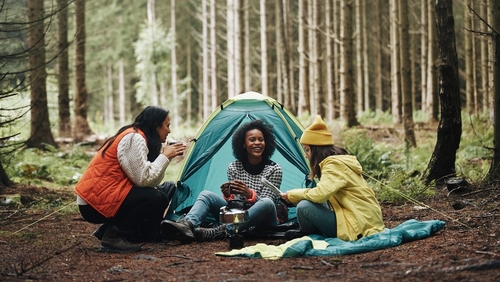 Three women are surrounded by woods and sitting around a small camping stove with a pot on top. There is a pitched tent in the middle, where one woman is sitting. On the left is a woman holding a cup with both hands and on the right a woman is sitting holding a piece of paper.