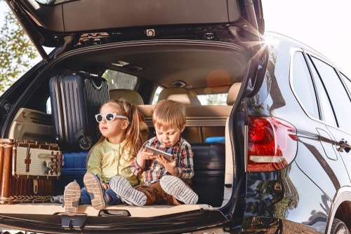 A boy and girl sitting in the trunk of car next to suitcases. Boy playing on tabley.