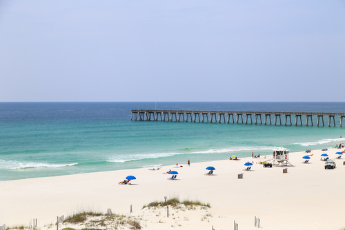 People relaxing and sunbathing on the beach with beach chairs and sunshades. In the back the Pensacola Beach Gulf Pier.