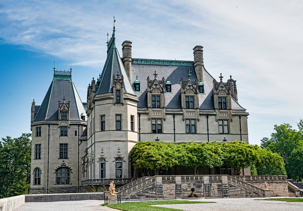 The historic Biltmore mansion is a mix of architectural styles but resembles a European chateau, built with large biege-gray stone and dark blue tiles on tall slanted roofs. Trees cover the front the mansion's veranda.