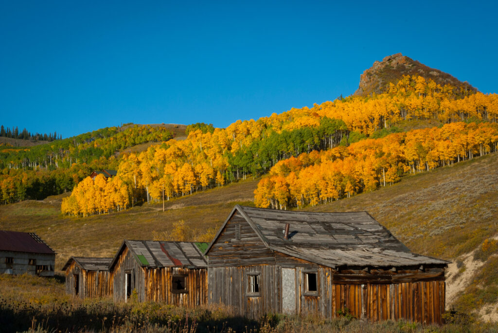 Several old wooden houses, with a background of rolling hills topped with trees with green and vibrant yellow leaves.