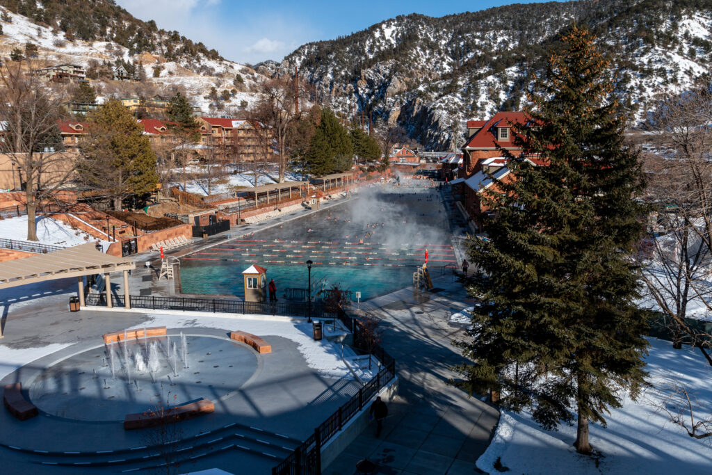 Glenwood Hot Springs pool with steam rising off the top, flanked by buildings, with snow-covered mountains in the background.
