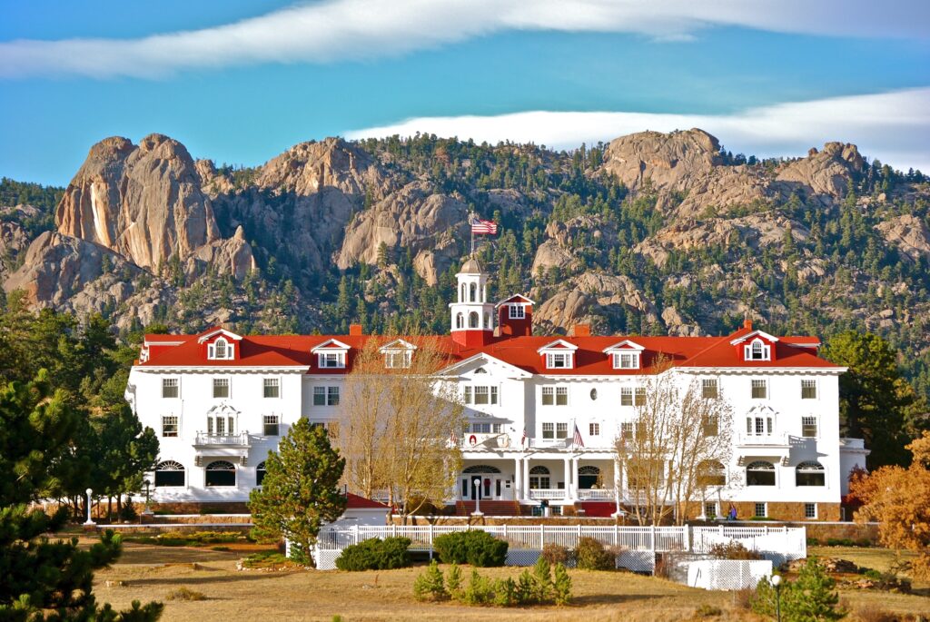 The Stanley Hotel, with its white façade and red-orange roof, against a backdrop of mountains dotted with trees.