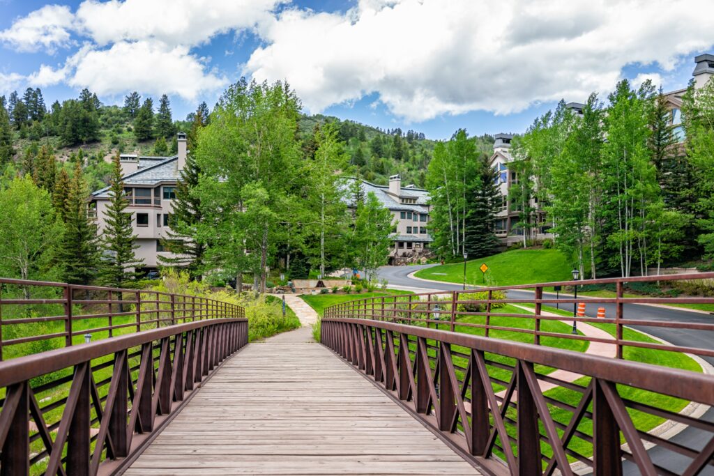 A wooden walkway with metal railings on either side leads to a neighborhood with buildings and many trees.