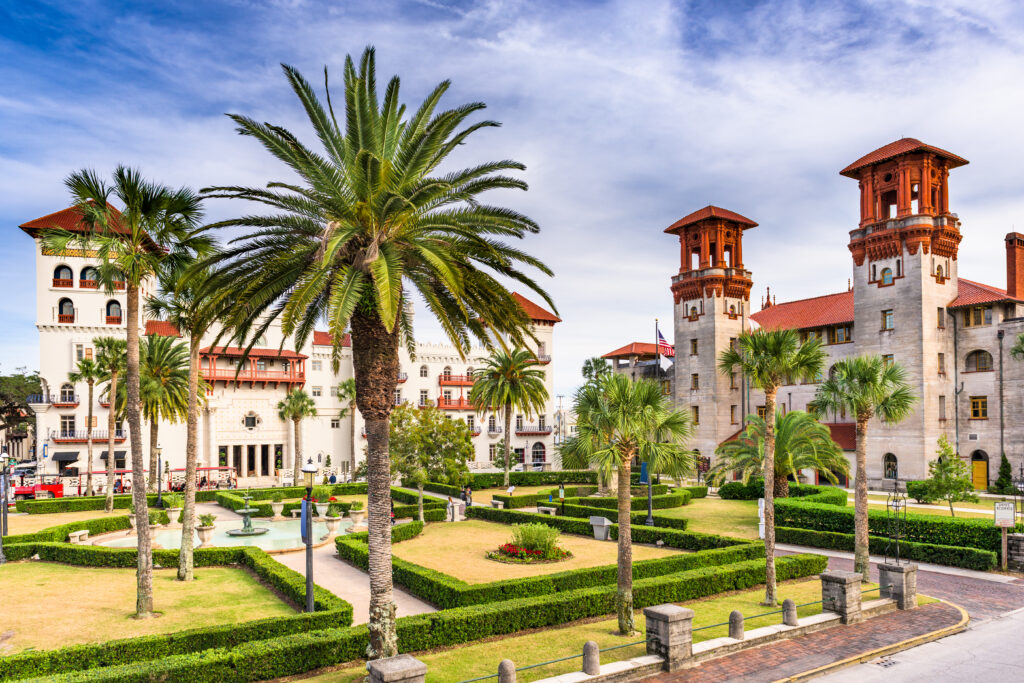 Buildings with European architectural influences have an off-white body and rust-red roofs, railings and balconies. In the foreground is a manicured garden with hedges, sculptures, walkways and palm trees.