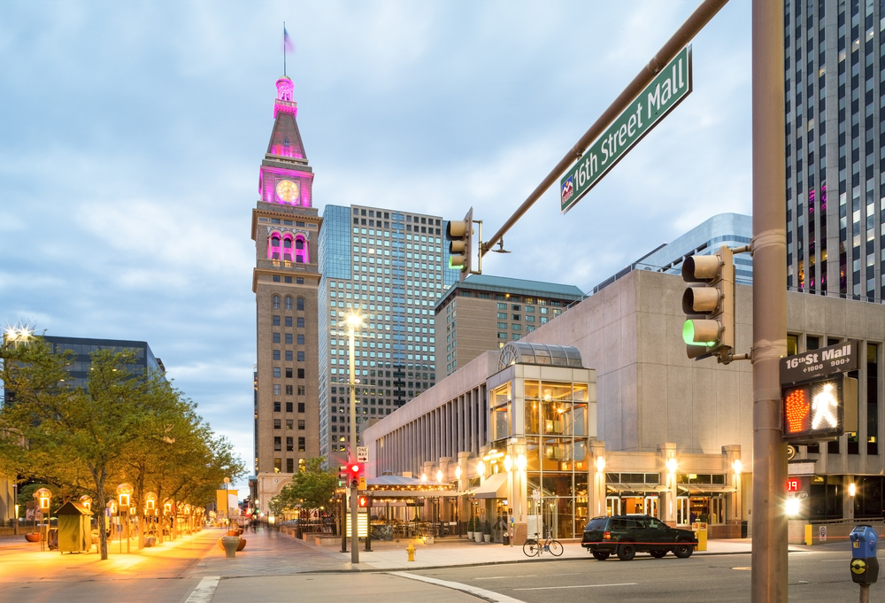 Street view of 16th Street Mall buildings and pedestrian path in Denver, with a clock tower in the background, lit at the top in pink lights.