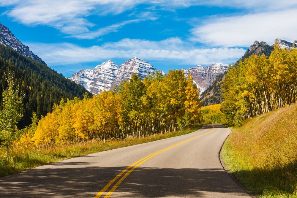 Scenic view along a road flanked with yellow and green trees, snaking toward snow-capped mountains