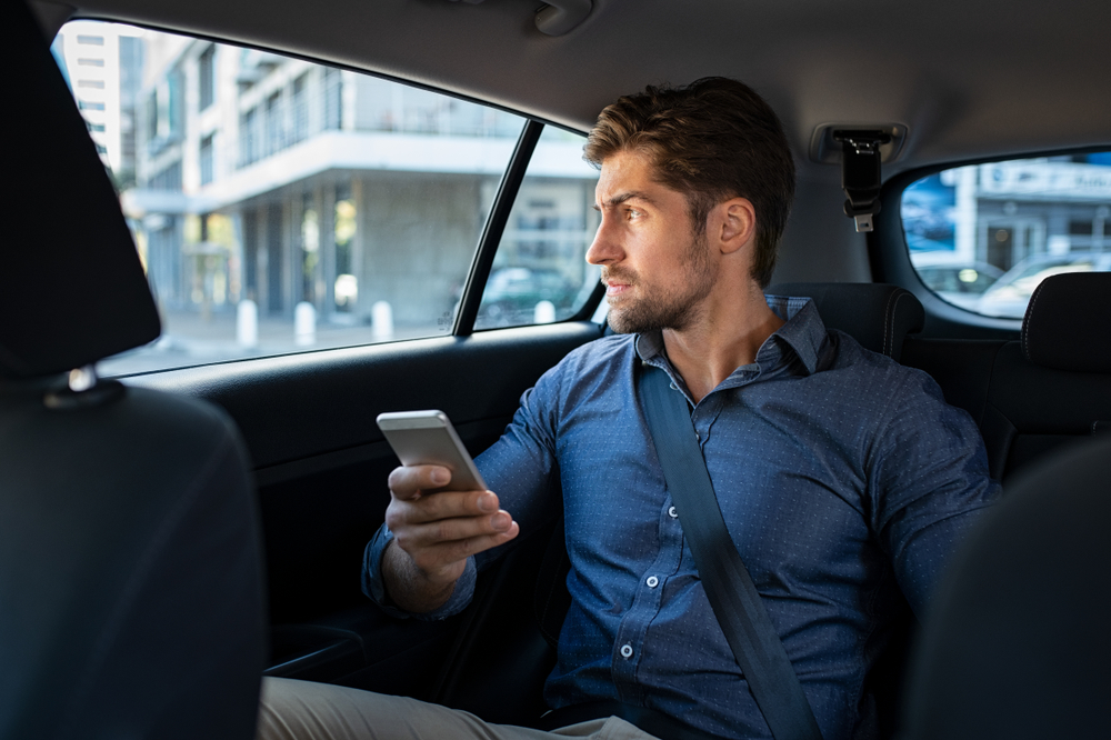 Thoughtful business man holding phone sitting in car looking out of window with serious expression.