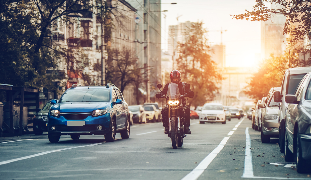 Motorcycle and cars are riding on a city street at the sunset.