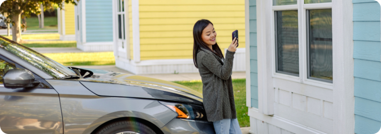 Woman leans against the hood of her car, looking at her cellphone and smiling.