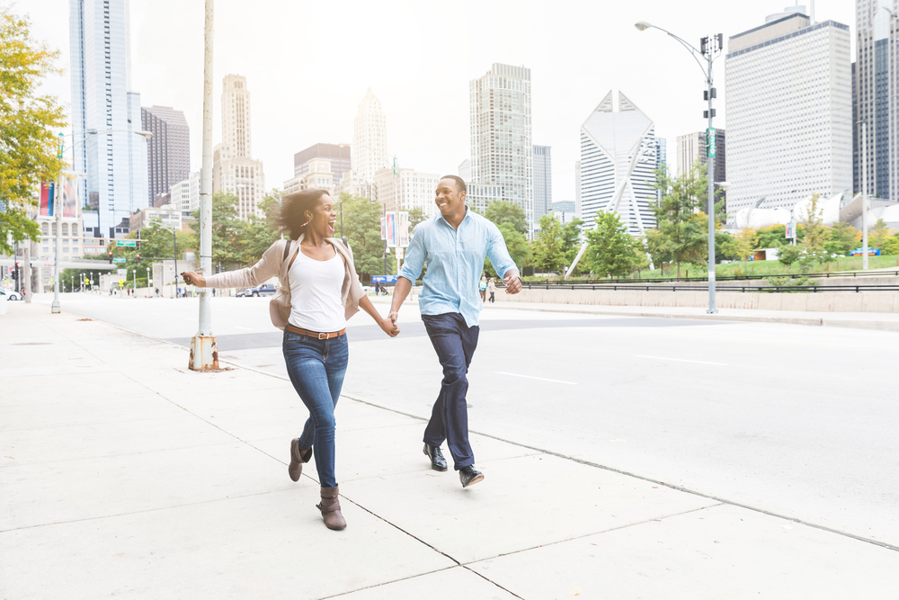 A man and a woman holding hands and running in the city. They are laughing and looking each other.