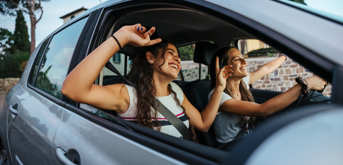 Two girls smile and dance along to a song while driving in a car.