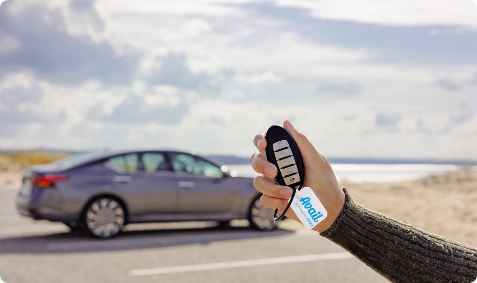 Hands hold car keys with an Avail keychain, with a car and beachscape in the background.