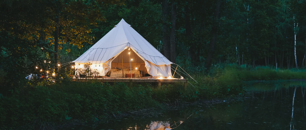 White tent illuminated with string lights sits on the bank of a river at dusk.