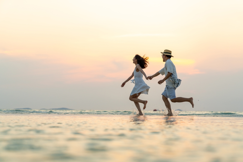 Happy couple holding hands and walking together on tropical beach at summer sunset on beach vacation.