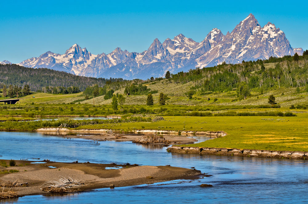 Grand Teton mountain range, partly covered in snow, with green forest, field, and small river in the foreground.