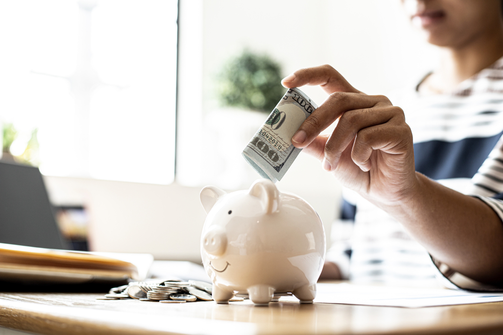 A business woman is putting dollar bills in a white pig piggy bank to save, she is doing an income and expense account to organize her financial plan.